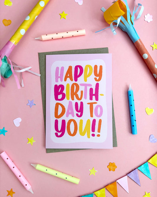 A colourful 'Happy Birthday to You!' greeting card with hand-lettered text in pink, orange, yellow, and purple, placed on a pink background with party props including candles, paper stars, and bunting.