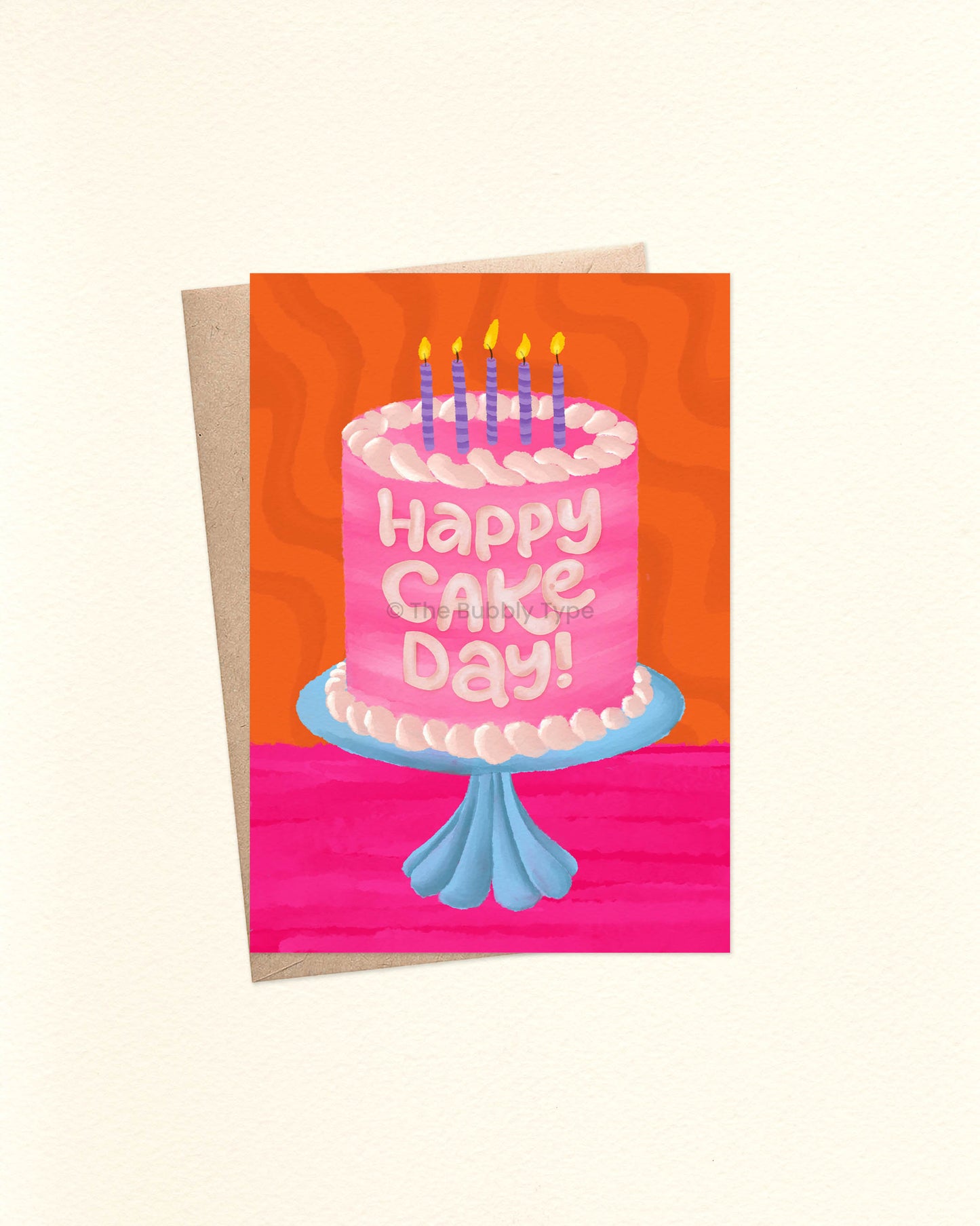 Front of an A6 birthday card featuring a watercolor illustration of a pink cake with pale pink-to-white frosting, sitting on a blue glass cake stand. The cake is topped with purple striped candles lit with yellow flames. The background is a vibrant hot pink and orange with a groovy wavy texture. The card is laid over a brown craft envelope.