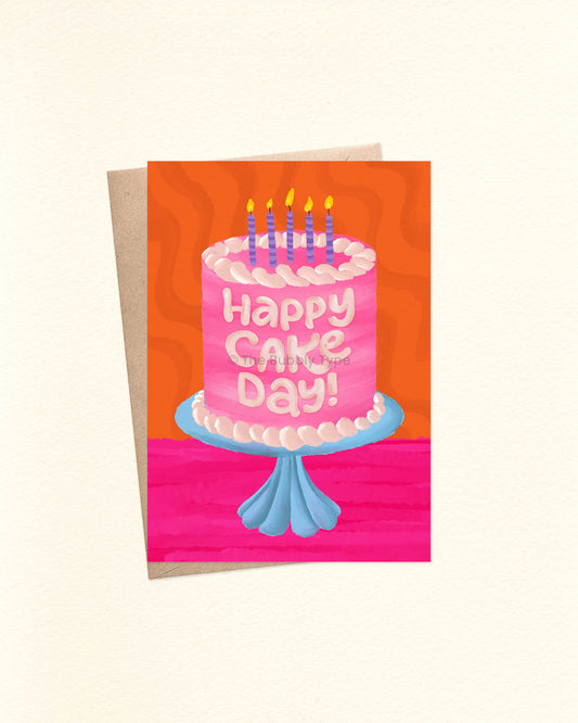 Front of an A6 birthday card featuring a watercolor illustration of a pink cake with pale pink-to-white frosting, sitting on a blue glass cake stand. The cake is topped with purple striped candles lit with yellow flames. The background is a vibrant hot pink and orange with a groovy wavy texture. The card is laid over a brown craft envelope.