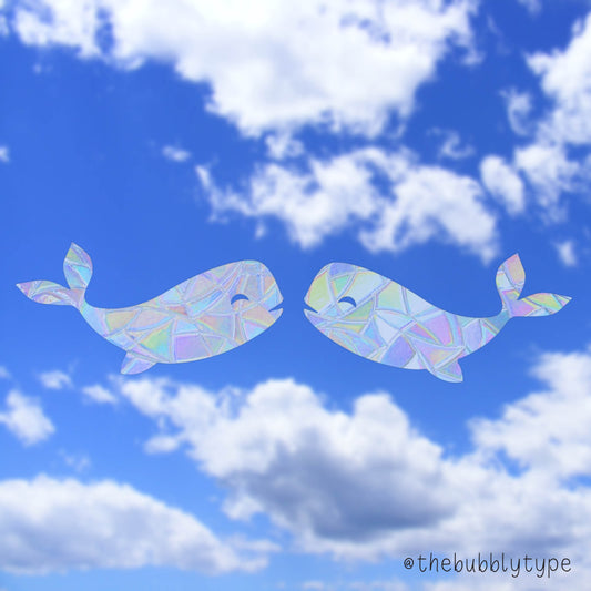Whale-shaped suncatchers facing each other, stuck to a window with a dramatic, cloudy blue sky as the backdrop—creating a peaceful, whimsical atmosphere. Kissing Whales look so cute in your window. Rainbow light reflections across the room. Looks great next to plants and crystals. Perfect home decor and makes the best gift for children and adults on their birthday or on Christmas.