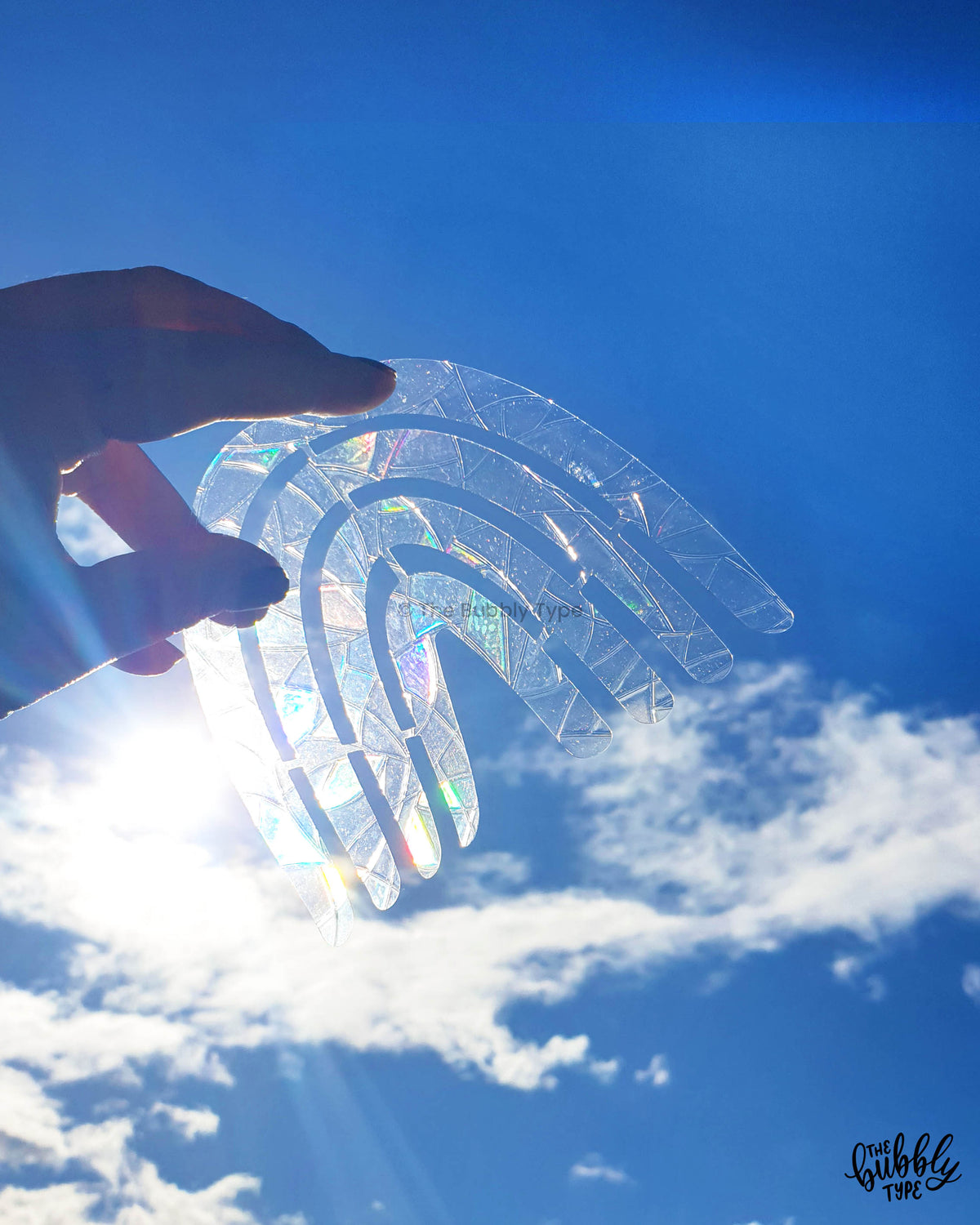Hand holding a rainbow arch sun catcher made from iridescent PVC cling film, catching the light with colourful reflections. Handmade Australian sun catcher gift by The Bubbly Type, perfect for windows, car mirrors, or as a small uplifting present.