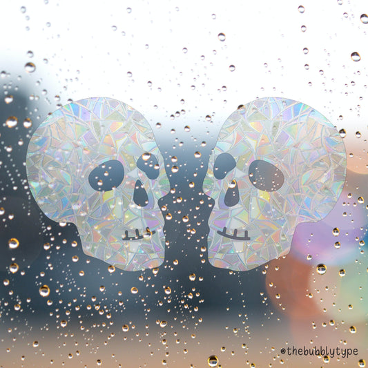 Two skull-shaped sun catchers facing each other on a wet window pane covered in raindrops. Behind them is a grey, gloomy blur with soft flares of colour from distant lights, possibly car headlights in the rain.