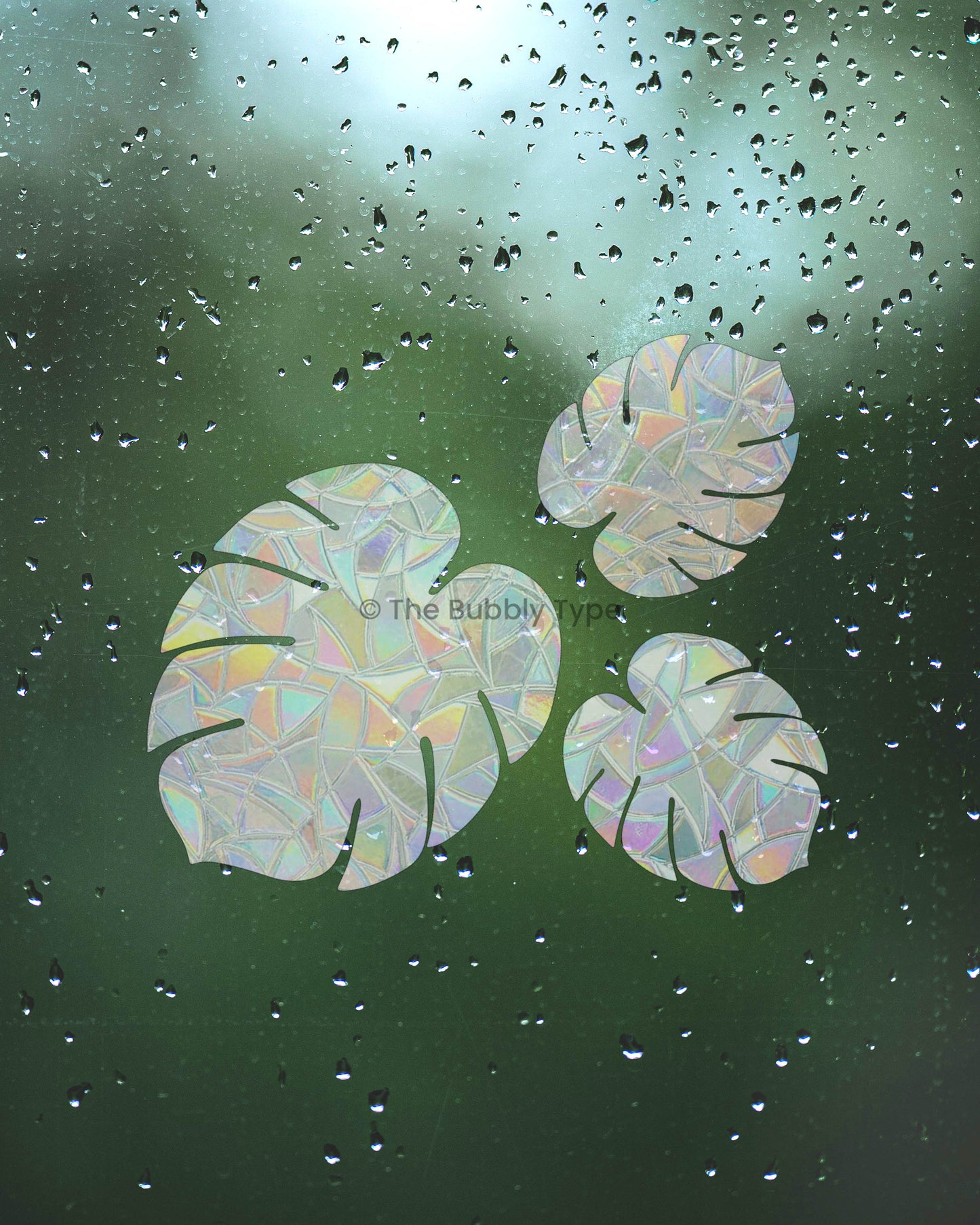 Three monstera leaf-shaped sun catchers facing each other on a wet window pane covered in raindrops. Behind them is a green blur of foliage with bright sunlight peeking through. The 3 monstera leaves are arranged to decorate your windows and make rainbows shine through when sun hits.