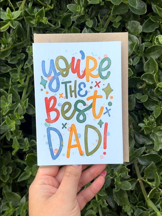 A hand holds a light blue greeting card that reads 'you're the best DAD!' in colorful playful lettering, with a brown kraft envelope behind it and green foliage in the background.