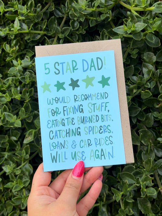 A hand with pink nail polish holds a light blue greeting card that reads '5 STAR DAD!' with star ratings and humorous phrases, in front of a background of green leafy plants.