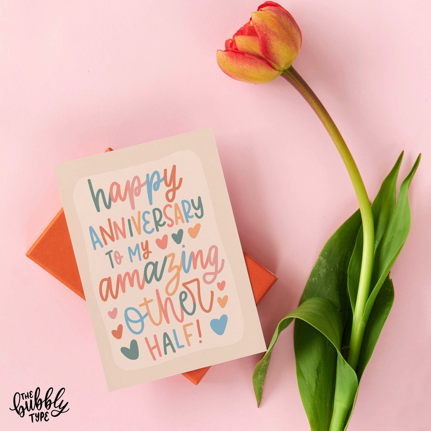 Australian made Love and Anniversary Greeting Cards. Happy anniversary to my amazing other half. Cute card for wife. Fun card for husband. Front of A6 anniversary card featuring whimsical cursive hand-lettering that reads: “happy anniversary to my amazing other half.” Lettering appears in soft neutral tones including dusky sage green, pale pink, peachy orange, and powder blue over a cream background. Shown with a tulip flower stem.