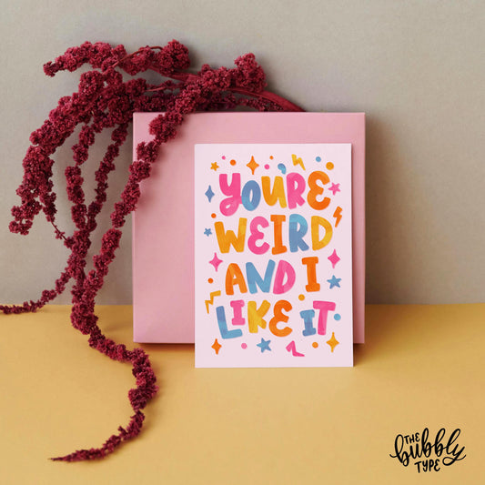 You're weird and I like it, A6 greeting card. Hand lettered design created by The Bubbly Type. The best vibrant Australian made cards you can find. Front of A6 greeting card with playful block hand-lettering that reads: “You’re weird and I like it.” The text appears in bright watercolour-style cutout letters in pink, blue, yellow, and orange on a pale pink background. Surrounded by colourful shape accents including stars, lightning bolts, dots, and squares.