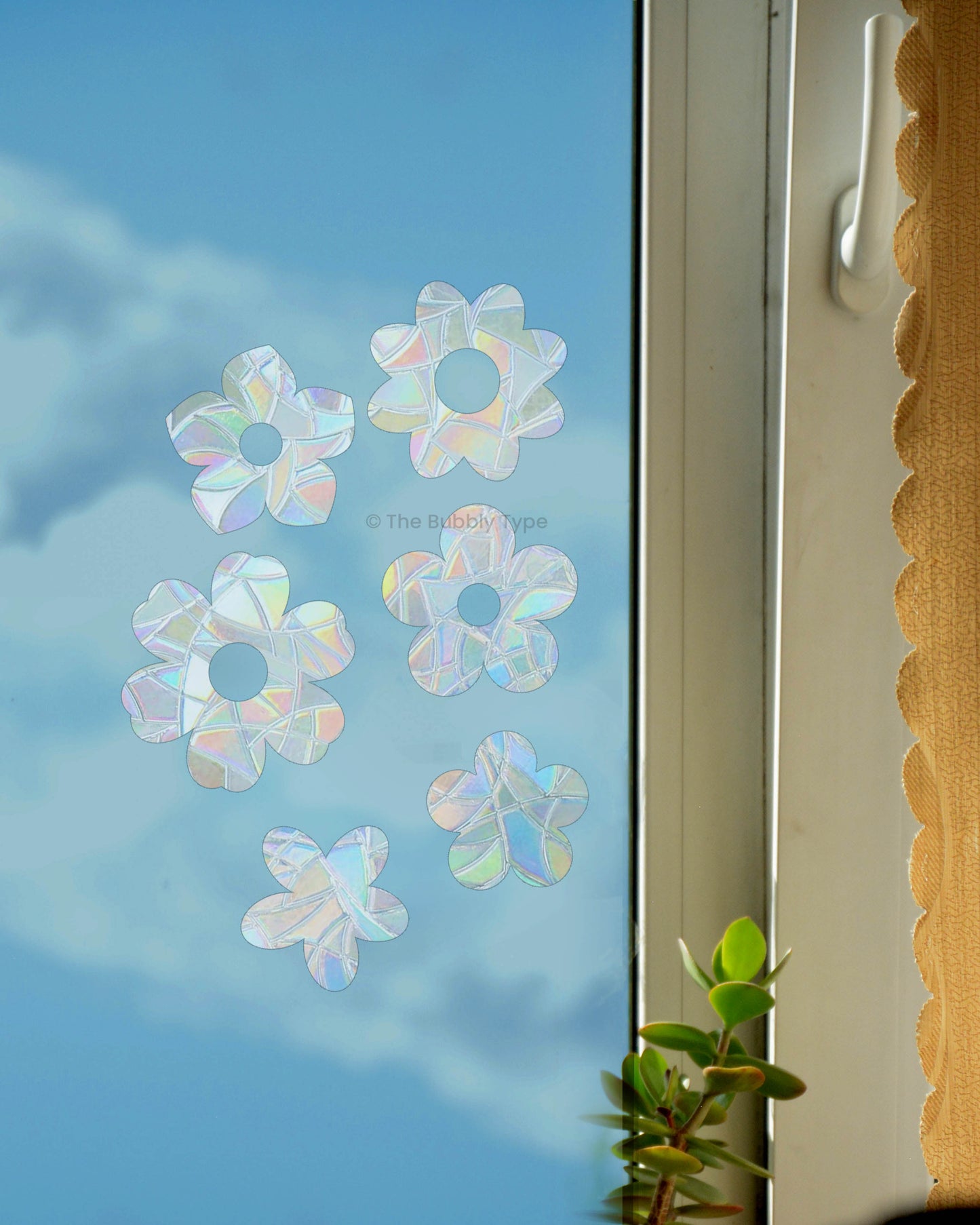 A range of flower shaped sun catchers stuck onto a window that shows blue skies. the white window frame and a peek of mustard curtains with scalloped edges is seen to the right. The sun catchers create rainbow light when the sun shines through them.