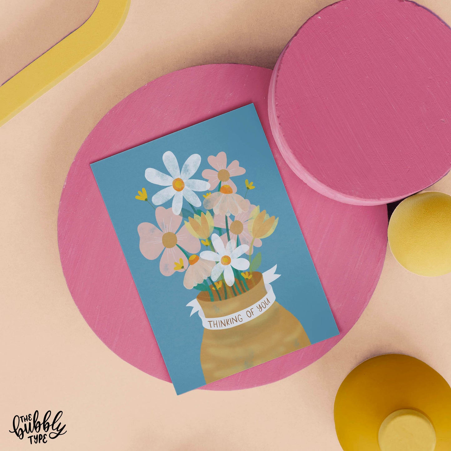 thinking of you greeting card with floral illustration made in Australia. best sympathy cards. A6 greeting card with a dusty blue background. The illustration features a mustrad coloured vase filled with pink, white and yellow flowers, and green foliage in a watercolour style. A white ribbon wraps around the neck of the vase with hand-lettered text reading “Thinking of You.”