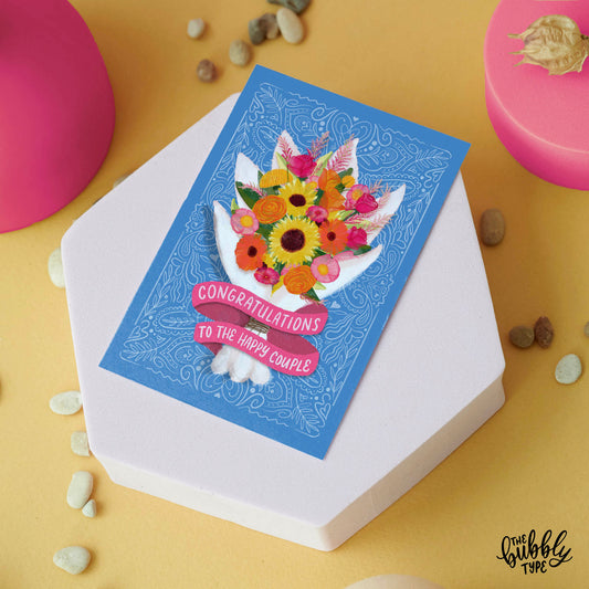 Australian made wedding cards and engagement cards. Congratulations to the happy couple bouquet of flowers. Front of A6 card designed for weddings or engagements. The background is a soft blue with fine white line pattern details. A vibrant watercolour-style bouquet takes centre stage, featuring sunflowers, hot pink and orange roses, pink peonies, yellow roses, and green foliage. The flowers are wrapped in a white paper wrap, with a pink ribbon banner at the bottom that reads: “Congratulations to the happy