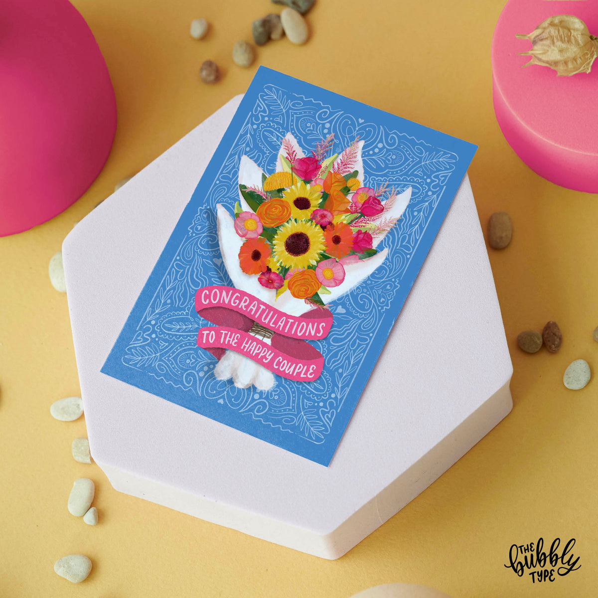 Australian made wedding cards and engagement cards. Congratulations to the happy couple bouquet of flowers. Front of A6 card designed for weddings or engagements. The background is a soft blue with fine white line pattern details. A vibrant watercolour-style bouquet takes centre stage, featuring sunflowers, hot pink and orange roses, pink peonies, yellow roses, and green foliage. The flowers are wrapped in a white paper wrap, with a pink ribbon banner at the bottom that reads: “Congratulations to the happy