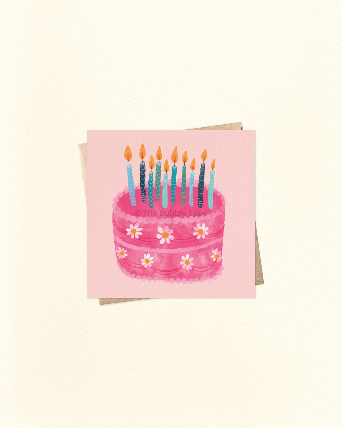 Front of 10cm square birthday card with a watercolour illustration of a pink birthday cake decorated with white daisy flowers and topped with multiple blue and teal candles. Set on a soft pink background and paired with a brown kraft envelope. A sweet and whimsical Australian made card by The Bubbly Type – perfect for birthdays of all ages.