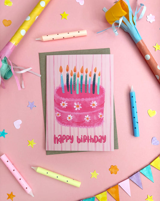 A pink birthday greeting card featuring a watercolour illustration of a pink cake with white daisies and lit candles, placed on a pink striped backing card. The card is styled in a flat-lay arrangement on a pink background with party props including colourful pencils, star confetti, paper hearts, bunting, and party hats.