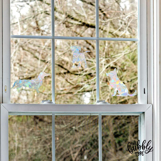 Three dog-shaped sun catchers — a dachshund, a Labrador puppy, and a German shepherd — displayed on a window with white horizontal and vertical bars. A bare tree with no leaves is visible outside through the glass. These doggy window stickers decals make rainbows when the sun shines through them. Made in Australia.