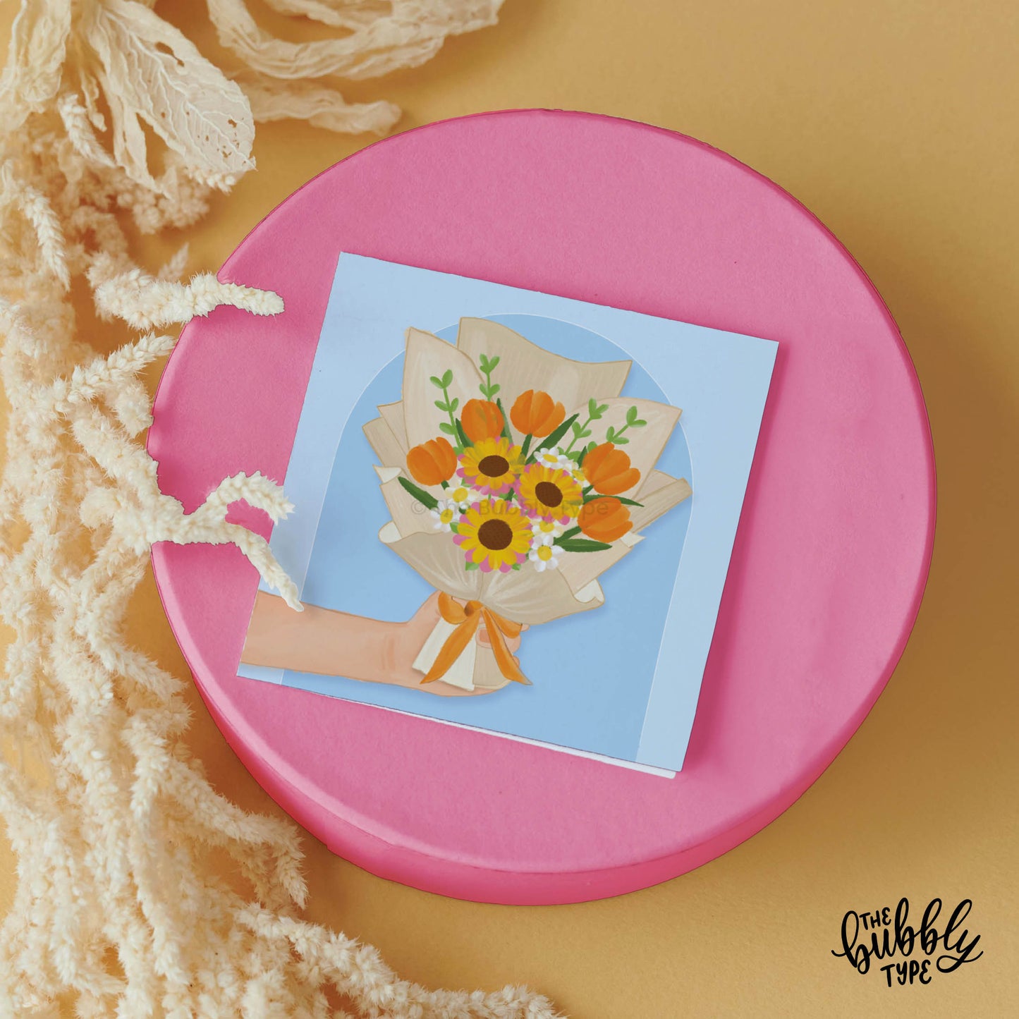 Front of 10cm square greeting card featuring an illustration of a hand holding a bouquet of orange flowers and other vibrant blooms, framed by an arch shape on a blue background. This card is shown lying on a circular pink platform with cream coloured dried flowers to the left.