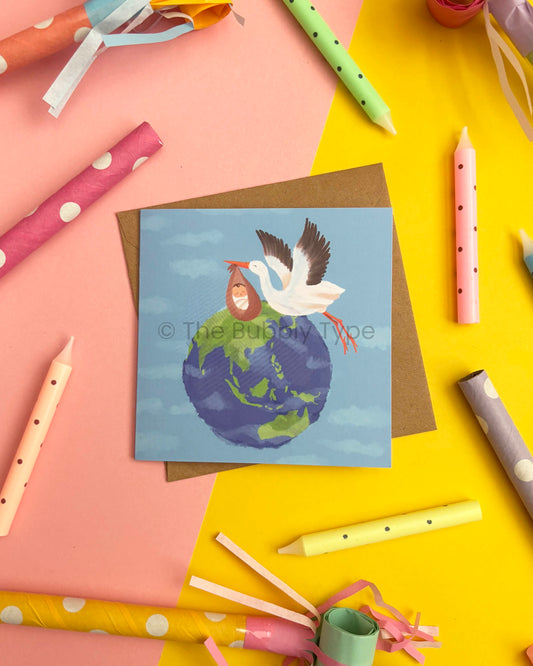 A square greeting card featuring an illustration of a stork carrying a baby in a bundle over a blue and green Earth, placed on a brown envelope. The print is photographed in a styled flat-lay with colourful candles and paper party horns on a pink and yellow background.