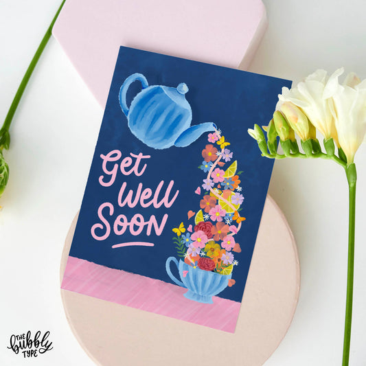 Best get well soon greeting card, made in Australia on 100% recycled paper. A6 greeting card with a rich deep blue background. The illustration shows a watercolor-style blue teapot tilted and pouring out a flow of yellow lemons, pink and orange flowers, green foliage, and butterflies into a matching blue teacup. To the right, in pink hand-lettered script, it reads “get well soon.”