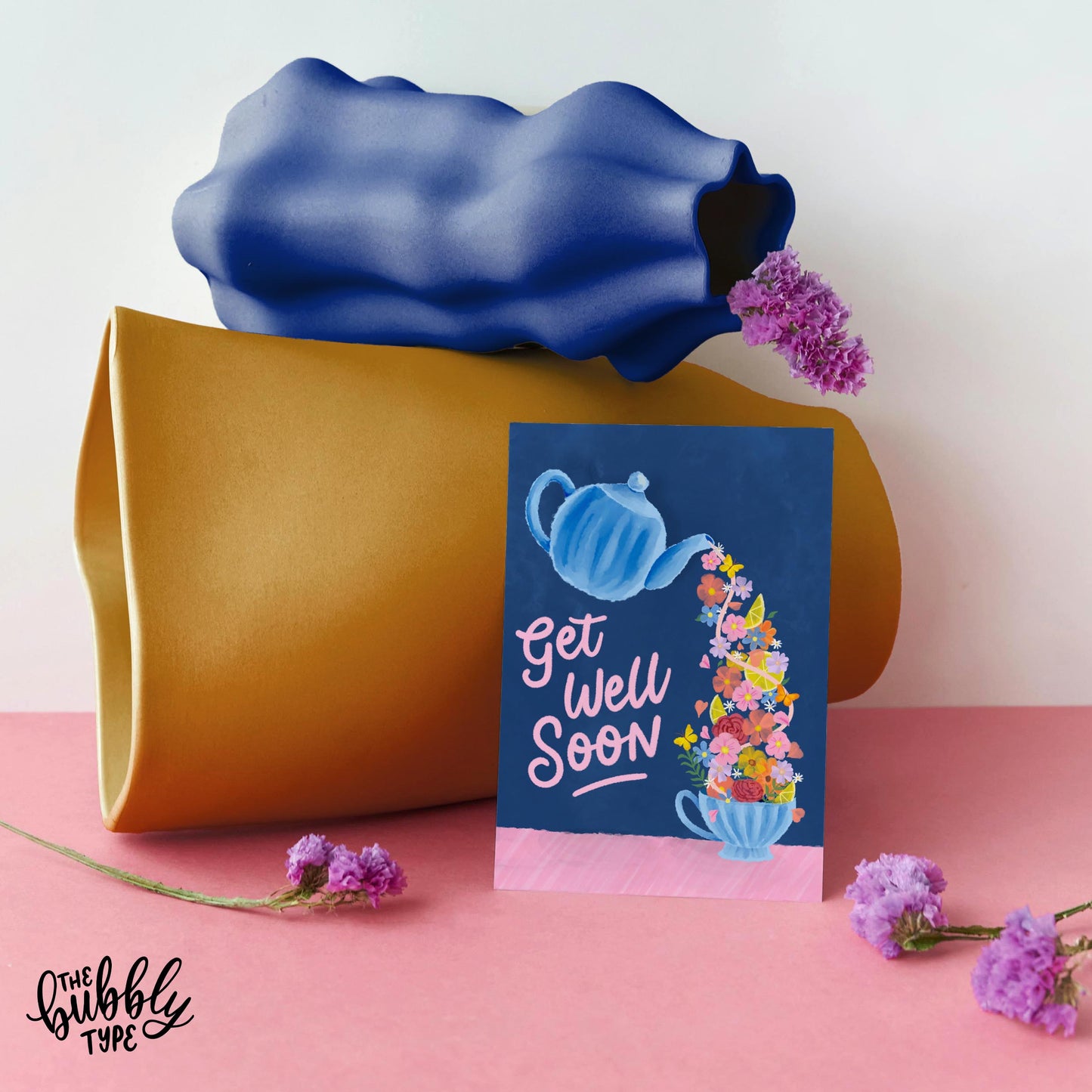 Get well soon card featuring an illustration of a teapot pouring flowers and lemons into a tea cup, itās perfect for conveying your heartfelt get well wishes. A6 greeting card with a rich deep blue background. The illustration shows a watercolor-style blue teapot tilted and pouring out a flow of yellow lemons, pink and orange flowers, green foliage, and butterflies into a matching blue teacup. To the right, in pink hand-lettered script, it reads āget well soon.ā