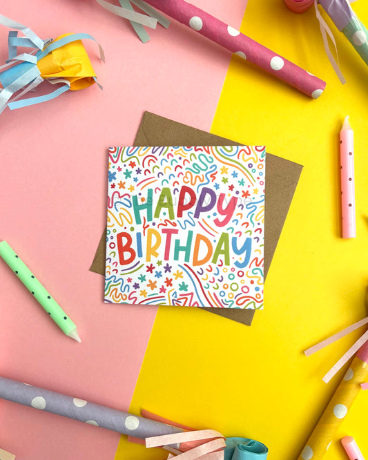 A square 'Happy Birthday' greeting card with colourful confetti and rainbow doodles, placed on top of a brown envelope. The card is photographed in a styled flat-lay with party props including pink and yellow paper party horns and pastel candles on a split pink and yellow background.