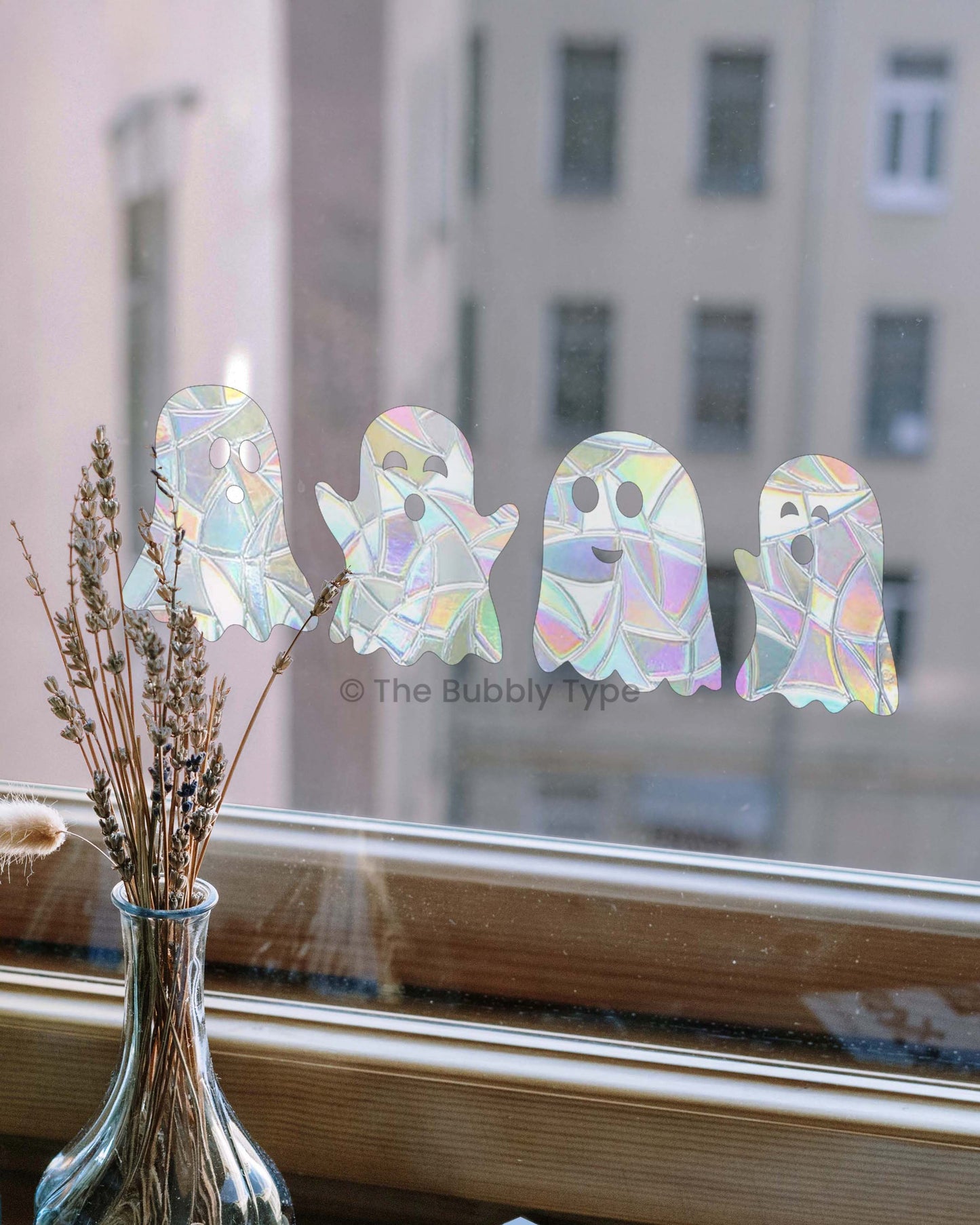 The same four ghost sun catchers arranged in a row on a window with a wooden sill. Each ghost shows a different facial expression, and the background reveals the windows of tall city buildings across the way.