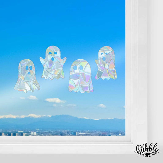 Four Halloween ghost-shaped sun catchers hanging in a white wood-framed window, with blue skies and soft clouds visible behind. Each ghost has a unique playful or spooky face expression, and the sunlight shines through their translucent designs.