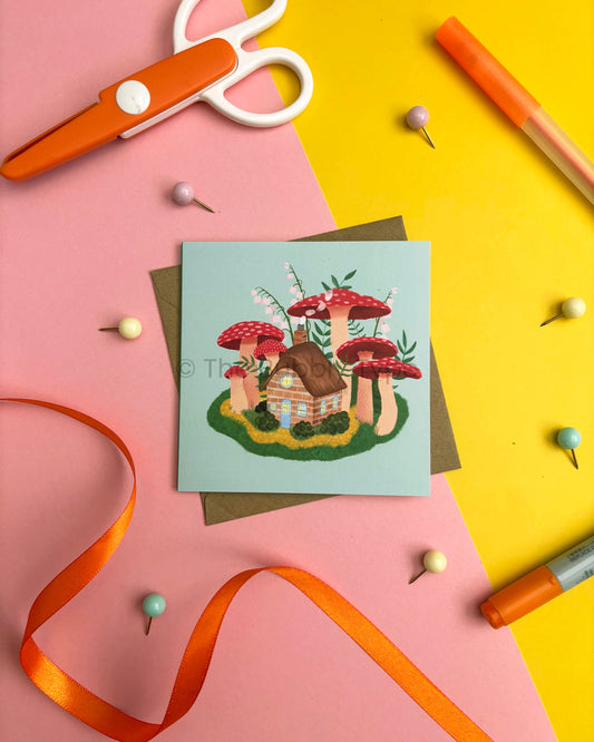 A square greeting card featuring a whimsical mushroom house illustration, styled in a flat-lay with an orange ribbon, scissors, pins, and markers on a split pink and yellow background.
