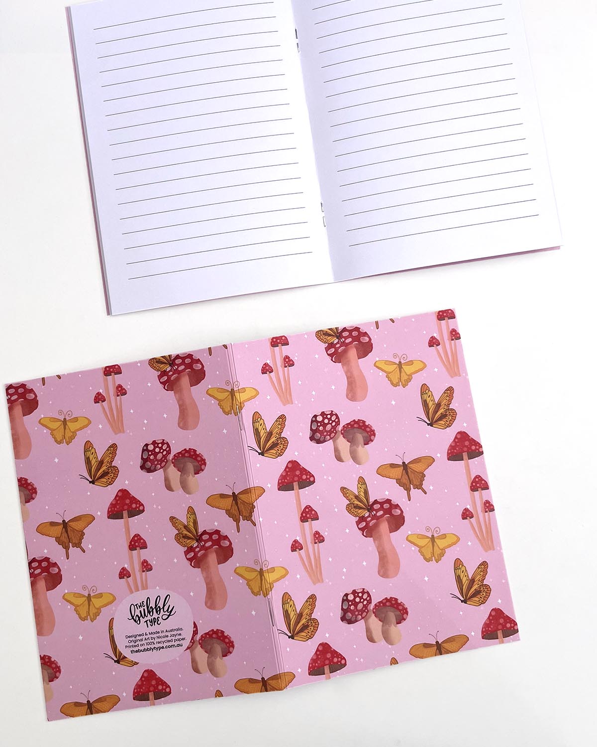 Two A5 Butterflies and Mushrooms notebooks. The top notebook is open to the middle, showing its white lined pages. The bottom one is open and facing down to display the back cover, which continues the butterfly and mushroom pattern. The Bubbly Type logo and print details are positioned at the bottom of the back cover.
