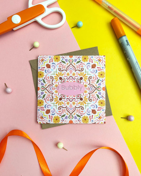 A square greeting card with a colourful floral mandala pattern and a central pink oval reading 'thank you', styled on a pink and yellow background with craft props including orange scissors, pins, a marker, and ribbon.