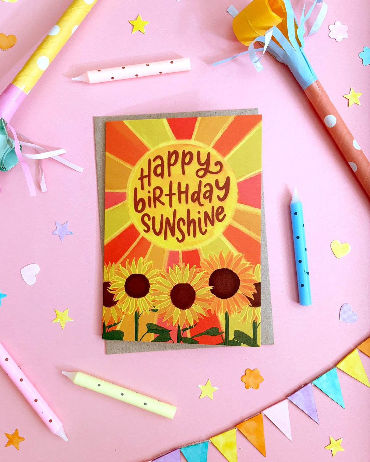 A birthday card with a sunburst design and three sunflowers, reading 'Happy Birthday Sunshine,' styled on a pink background with party candles, streamers, and star confetti.