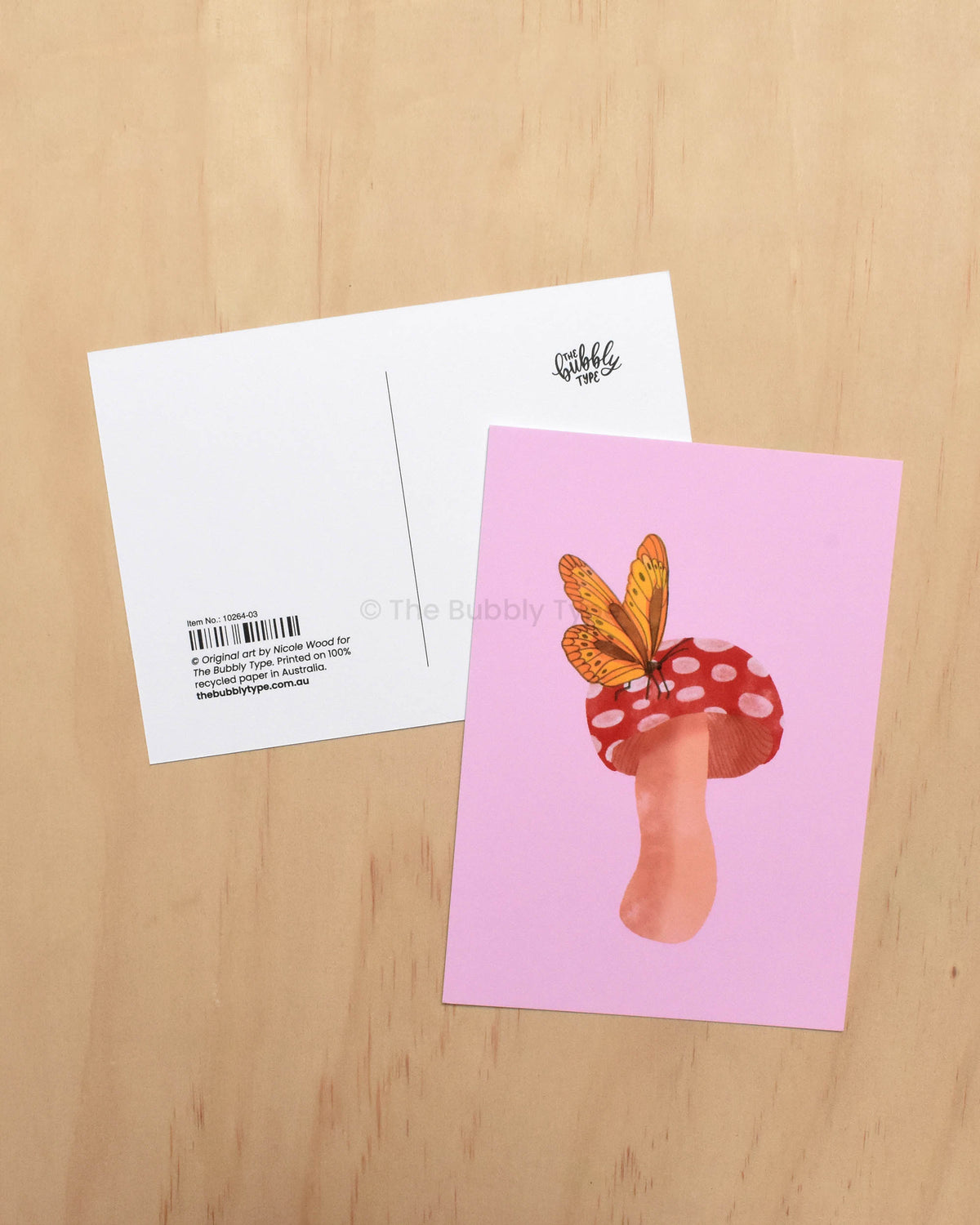 Postcard with a mushroom and butterfly design on a pink background, placed on a wooden surface.