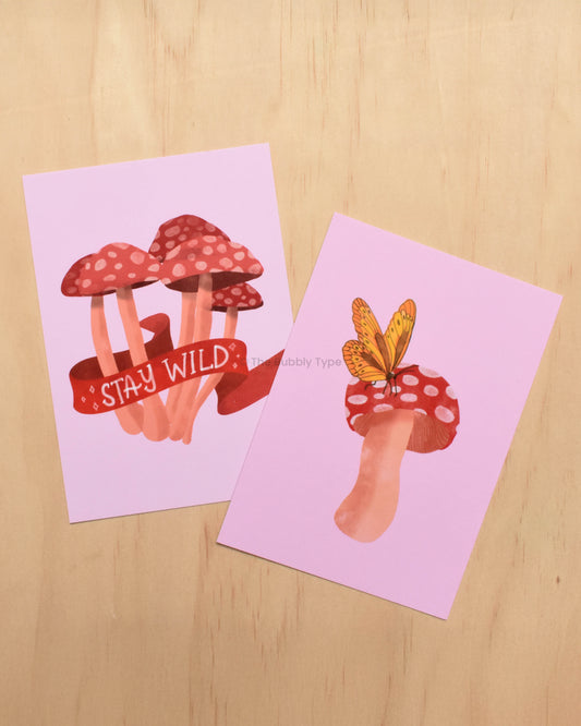 Two pink art postcards featuring wild mushroom and butterfly illustrations on a wooden background.