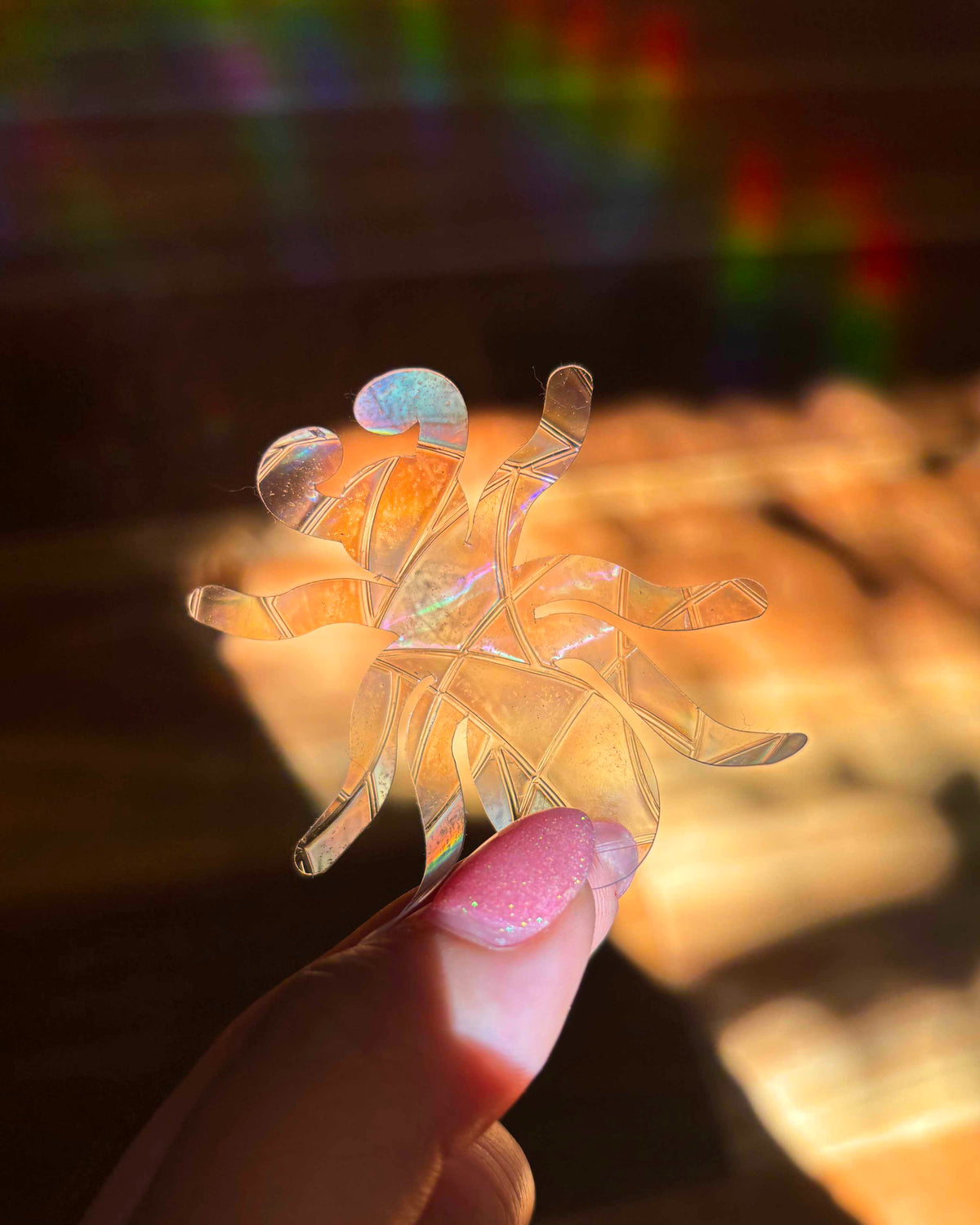 Clear plastic ant-shaped sun catcher held by a hand with blurred colourful rainbow lights in the background