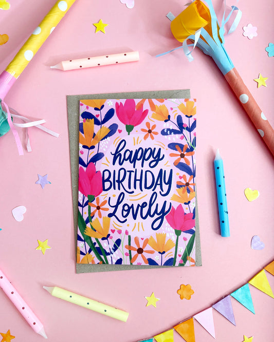A floral birthday card with the words 'happy BIRTHDAY Lovely' in navy script, surrounded by colourful party props on a pink background.