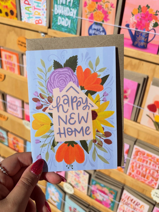Card with 'Happy New Home' text and floral design held in front of a display of greeting cards.