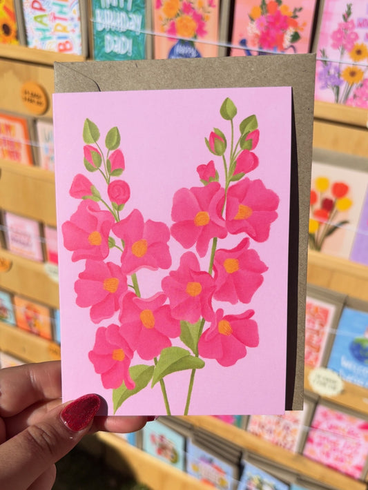 Hand holding a pink floral card with green leaves against a background of other greeting cards.