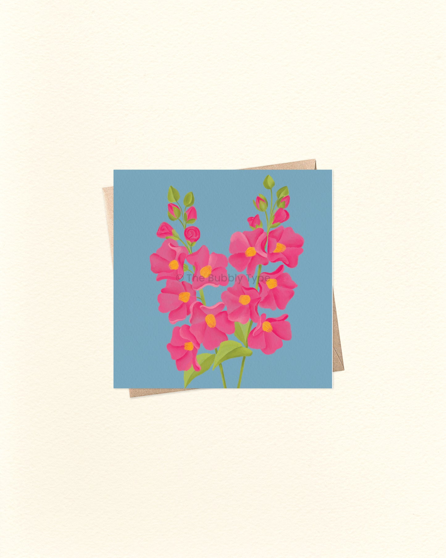 A square greeting card featuring a stylised floral bouquet in pink with yellow centres and green stems on a light blue background, shown with a kraft envelope behind it on a warm off-white surface.