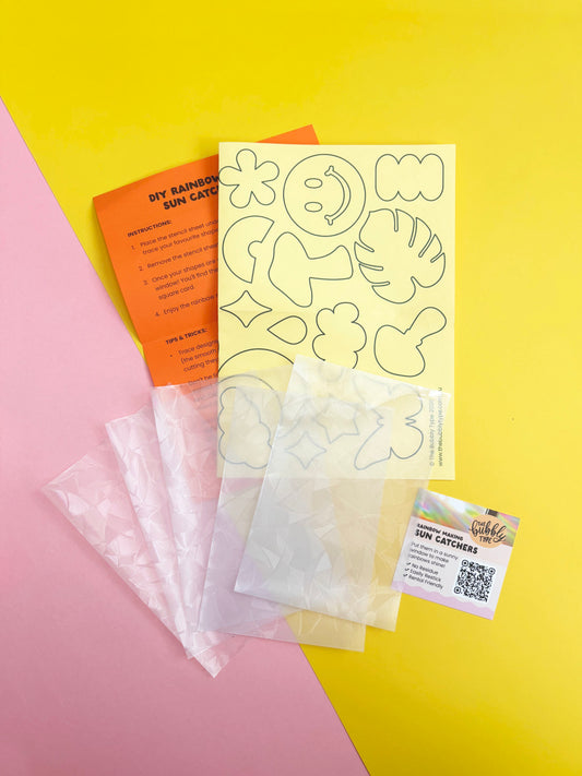 DIY rainbow sun catcher kit contents laid out on a diagonally split pink and yellow background, including clear sheets of sun catcher material, a yellow stencil sheet with outline templates, an orange DIY instruction card, and a small branded card with installation instructions.