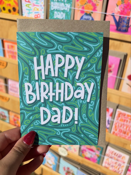 Hand holding a birthday card with 'Happy Birthday Dad!' text against a store background.
