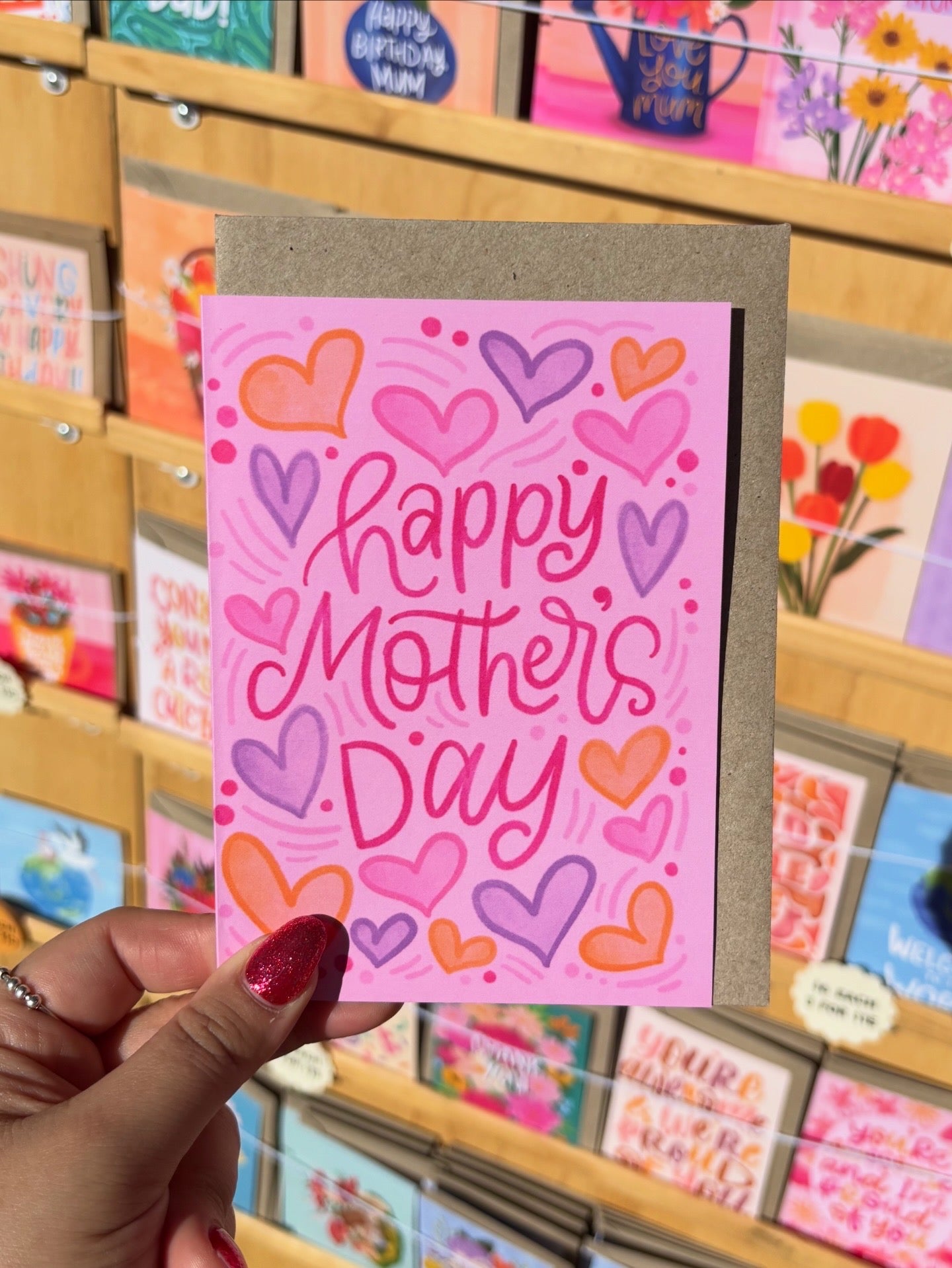 Hand holding a 'Happy Mother's Day' card with colourful hearts in front of a display of greeting cards.