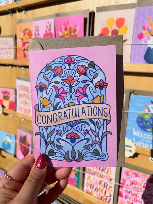 Hand holding a 'Congratulations' card with floral design in front of a display of other cards.