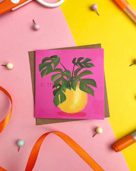 A square greeting card featuring a green monstera plant in a yellow pot against a bright pink background, styled on a brown envelope over a split pink and yellow surface with ribbon and pins.