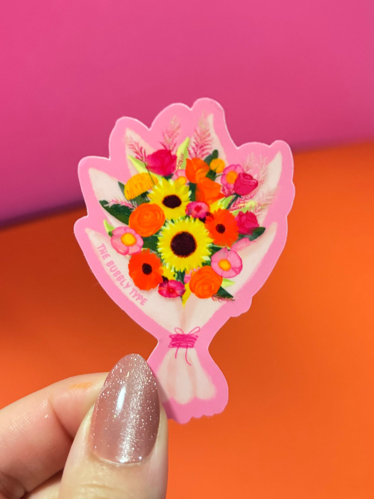 A hand with glittery pink nails holds a small pink flower-shaped sticker or charm featuring a colourful bouquet of sunflowers and orange flowers. The background is a bright, solid orange surface with a pink upper backdrop.