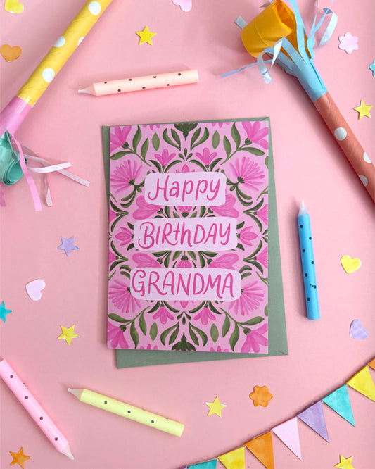 An Australian-made birthday card for 'Grandma' featuring a pink and green floral damask pattern with 'Happy Birthday Grandma' written in pink letters. The card is styled in a flat-lay arrangement on a pink background with party props like candles, paper party hats, and star confetti.