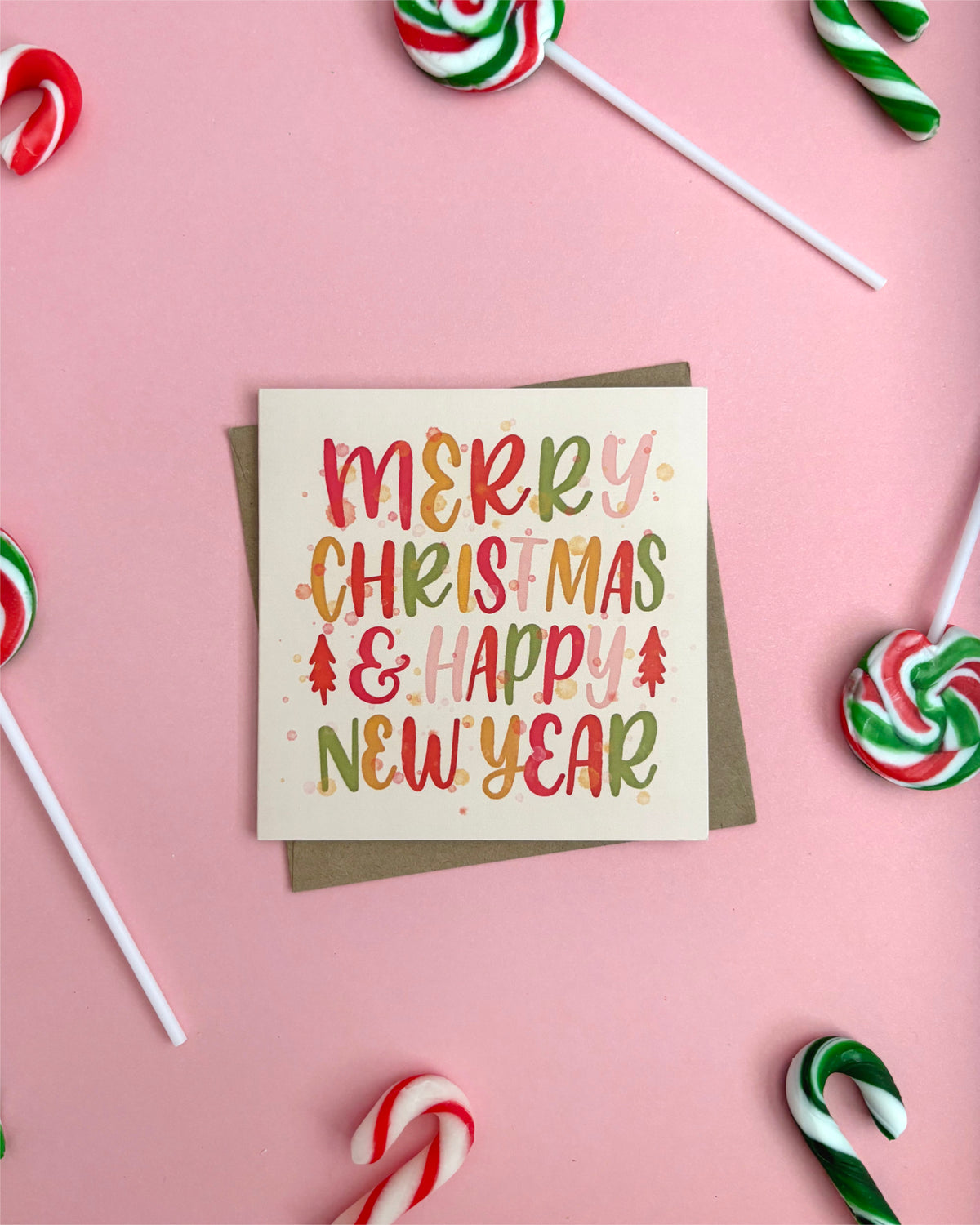 A square Christmas greeting card with a kraft envelope, placed on a pink background and surrounded by candy canes and lollipops. The card reads “MERRY CHRISTMAS & HAPPY NEW YEAR” in colorful hand-lettered text.