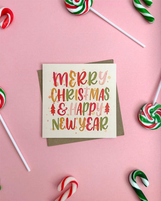 A square Christmas greeting card with a kraft envelope, placed on a pink background and surrounded by candy canes and lollipops. The card reads “MERRY CHRISTMAS & HAPPY NEW YEAR” in colorful hand-lettered text.