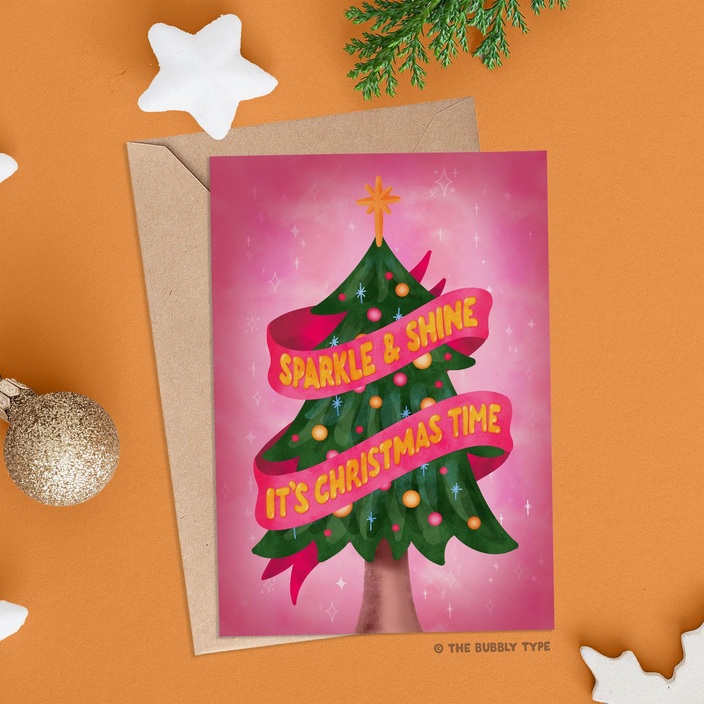 Card displayed on an orange background with a brown kraft envelope underneath. Surrounding the card are cheerful Christmas decorations, creating a festive, cozy vibe.
