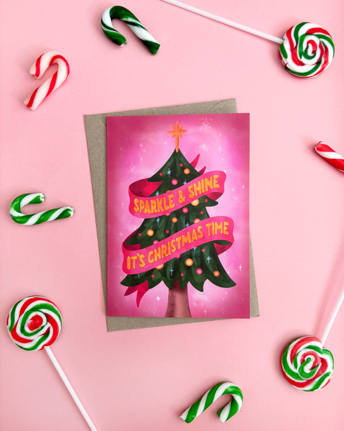 A Christmas greeting card featuring a stylized green Christmas tree with pink ribbons and the text 'SPARKLE & SHINE IT'S CHRISTMAS TIME' on a pink background, styled on a pink surface with candy canes and lollipops around it.