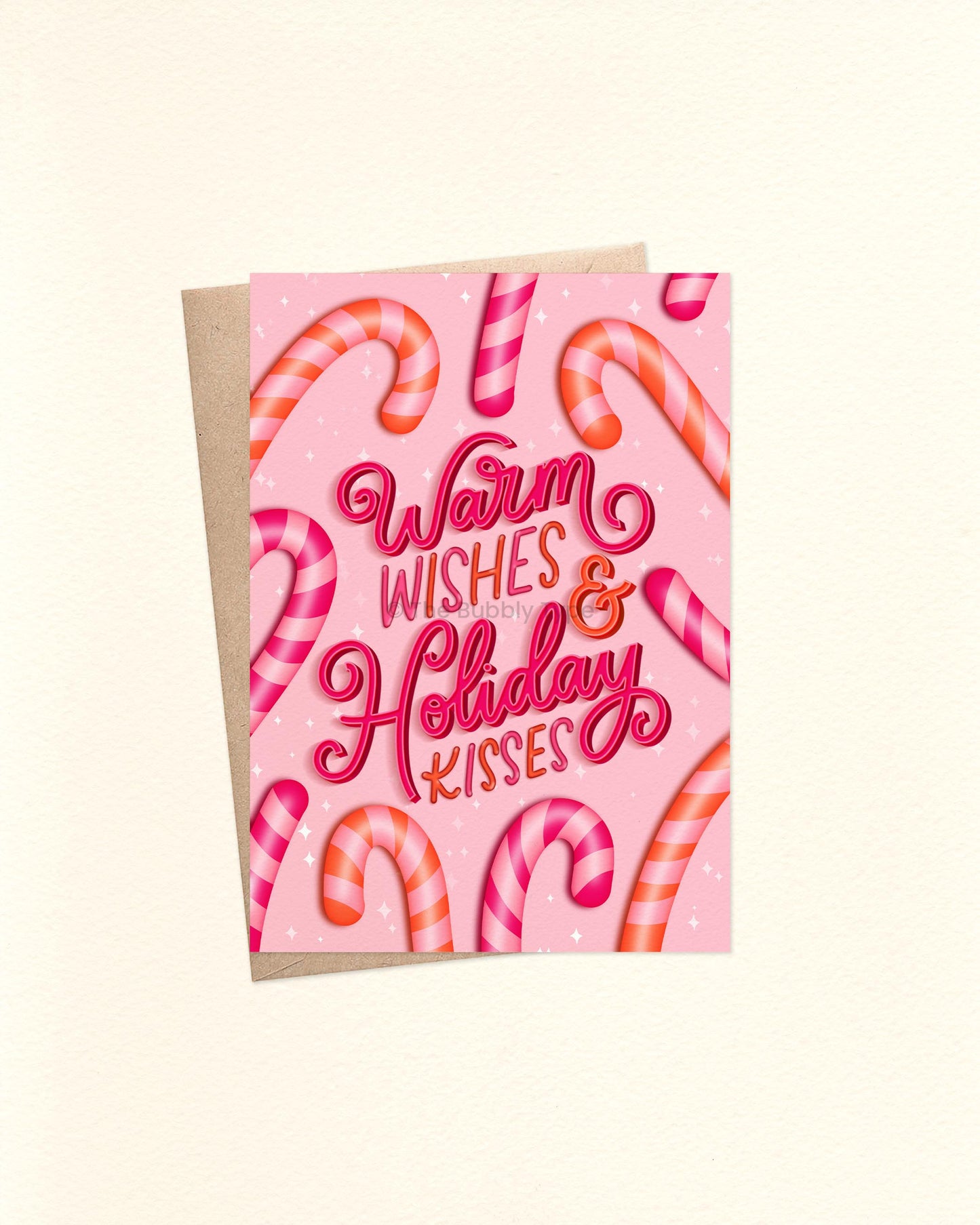 A6 Christmas card featuring a playful pink background with an illustrated border of pink and orange candy canes. In the center, bold hand lettering in hot pink and orange reads “Warm Wishes and Holiday Kisses.” The card is laid flat with a brown kraft envelope underneath.