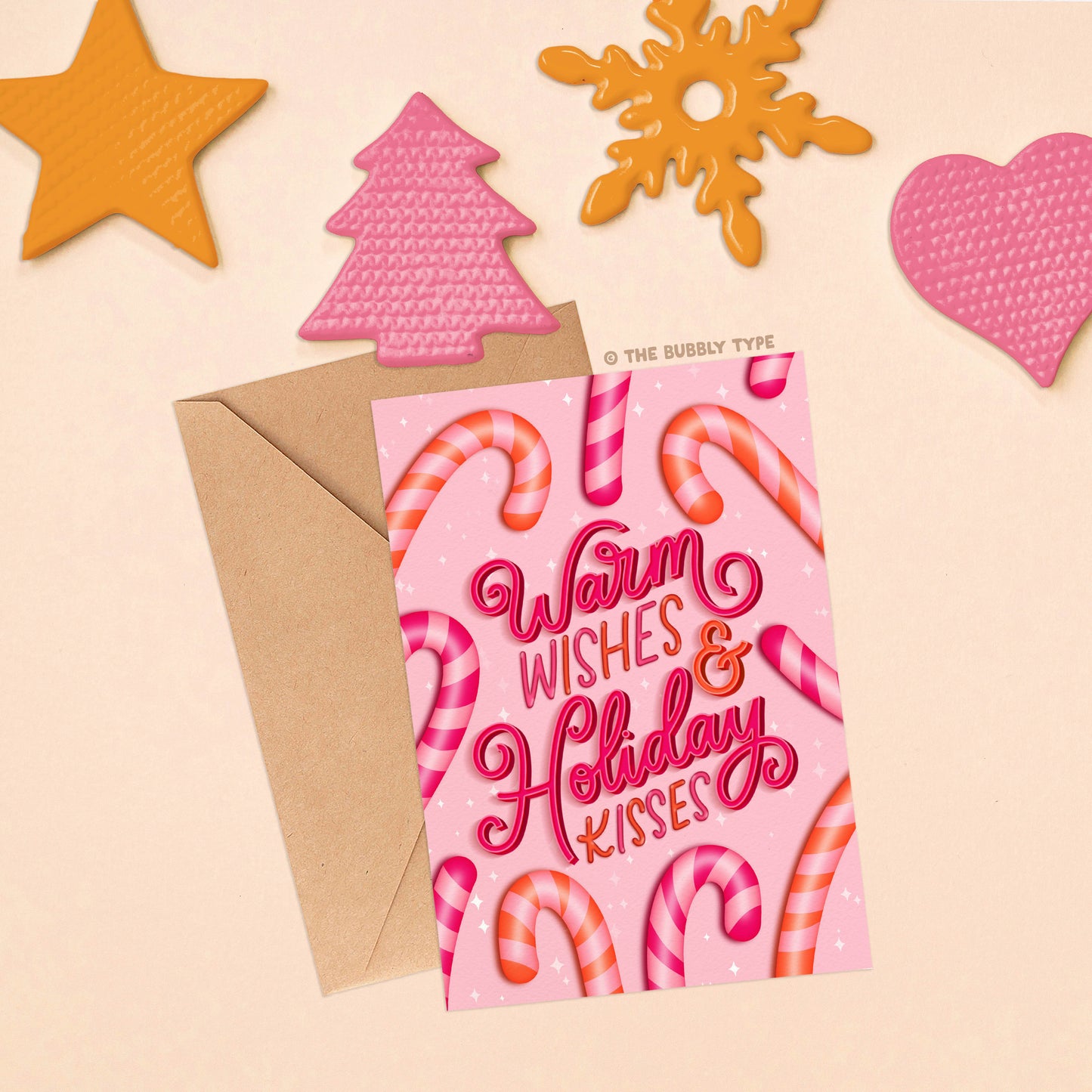 Card and envelope laid flat on a soft background with a festive arrangement of Christmas ornaments in orange and pink at the top, including shapes like a star, Christmas tree, snowflake, and heart.