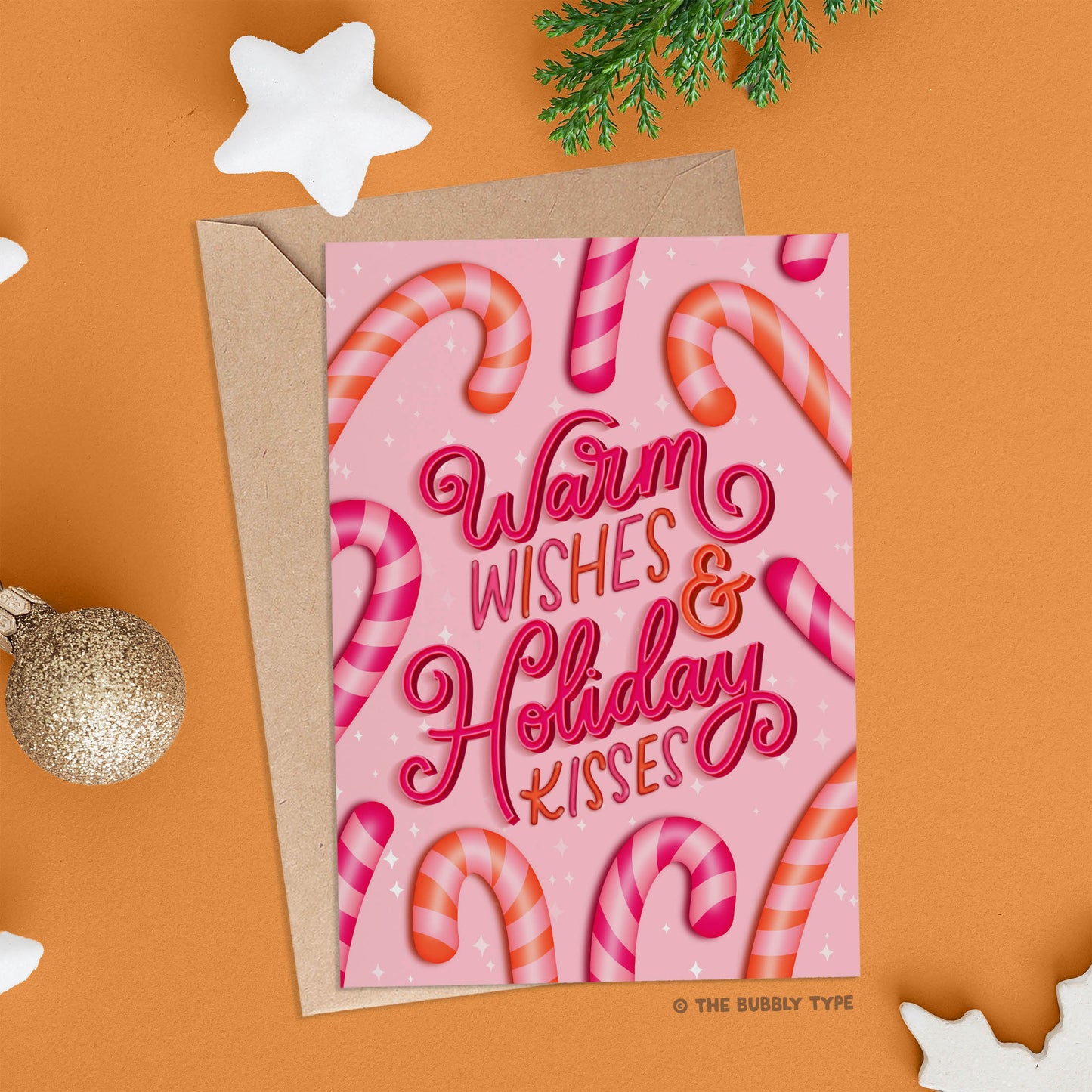 The same Christmas card shown on an orange background, laid flat with its kraft envelope. Surrounding the card are festive decorations including a gold glitter bauble and white glitter star ornaments.